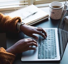 person typing on laptop next to coffee mug and notebook with pen