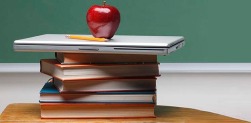 books stacked on a desk with a laptop and apple on top in front of a chalkboard